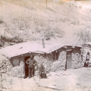 Miners pose near mud plastered dugout houses (a duplex) in Colorado. they wear hats with miners lights. One man smokes a pipe.