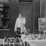 A man smokes and stands near his display of "Santa Fe Pottery" on a sidewalk at the Cinco de Mayo Festival hosted by the West Side Coalition on Santa Fe Drive in the Lincoln Park neighborhood,  Denver, Colorado. The pottery is displayed on tables covered with Mexican blankets and sheeting. A sign in a window reads: "New & Used Furniture."