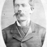 In This portrait of the miner nicknamed "Howling Hyena", taken in  Leadville, Colorado, he wears a coat with short collar lapels and a cravat with a stiff collar. His hair is thick and neatly combed and he has a long mustache with a small chinwisker below his lower lip.