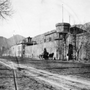 Stone walls of the State Penitentiary in Canon City, Colorado, line the sidewalk. A horse drawn water wagon on the dirt street is in front of a faceted tower with a balcony. A stone gate next to it is carved with entablature, and the original administration building is set back.