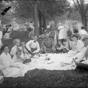 Outdoor portrait of men, women, girls, boys, and teenagers at a picnic in a Denver, Colorado park; costume includes a large hats with flowers. A man in military uniform eats a banana; a boy holds a a United States flag.