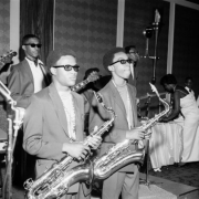 Members of a teenage jazz band stand on a stage at a dance in Denver, Colorado. Some of the African American (Black) musicians have their hair styled in short Afros and wear thick framed black glasses. The saxophone players wear turtlenecks, suit jackets, and thick gold chains. Shows saxophones, base guitar, lead guitar, and drums. Women in white full length formal dresses and men in tuxedos are in the distance.