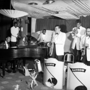 View of a Futuristics Club function. Members of an orchestra play on the stage of the Rainbow Ballroom, 38 E. Fifth Avenue (5th Ave.), Denver, Colorado. The African American (Black) band members play the piano, saxophone, trumpet, drums, and bass. They wear white jackets and stand in front of music stands that read: "Rainbow." Another sign reads: "No Smoking on Orchestra Stand at Any Time - Management."