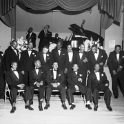 View of an Idle 12 Club function. A group of African American (Black) men pose in front of a stage at a dance hall in Denver, Colorado. The men wear tuxedos and bow ties. A jazz band is on the stage in the distance. On the wall near the stage a sign reads "Stag Show, Feb. 1955."