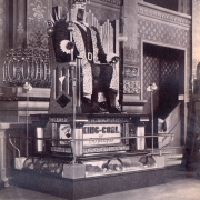 View of the statue of King-Coal against a backdrop of gilded plasterwork and ornate wallpaper in the Colorado Mineral Palace, Pueblo (Pueblo County), Colorado. The statue is made of coal, diamonds, and diamond dust, the figure is seated on a pedestal of copper and nickel with a glass display case below. Illumination is supplied by four dual-bulb floor lamps, one at each corner of the pedestal. A sign reads: "King-Coal of Trinidad." Shows a stairway to an upper floor through an archway.