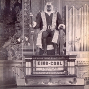 Close-up view of the statue of King-Coal in the Colorado Mineral Palace, Pueblo (Pueblo County), Colorado. The statue is made of coal, diamonds, and diamond dust, the figure is seated on a pedestal with a glass display case below. Lighting is supplied by four dual-bulb floor lamps, one at each corner of the pedestal. A sign reads: "King-Coal of Trinidad." Shows part of the stage with stalactite and stalagmite decoration. A pipe organ is nearby with a sign that reads: "The Pueblo Smelting [?]"