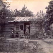 View of the oldest house in Salt Lake City (Salt Lake County), Utah; shows a one story hewn log cabin with chinking and a hewn board roof, a double horizontal window with panes of glass, a door and stone chimney. A man and two young boys stand and sit on chairs and benches near the house. Shows a small dirt yard with garden, a cold frame, a wooden push cart with one wheel, a wooden gate, trees. Shows part of a two story brick masonry house.