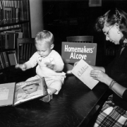 A woman reads a book titled "Living with Children" in the Homemakers' Alcove at the Denver Public Library at Colfax Avenue and Bannock Street in the Civic Center neighborhood of Denver, Colorado. Her baby boy sits on the table and plays with a bound volume of Parents' Magazine.