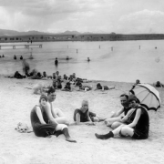 Young men and women sit on the beach at Berkeley Park in Denver, Colorado. The women wear bathing suits, bathing caps, and one woman rests a parasol over her shoulder. The men and boys nearby also wear bathing suits. Swimmers are in Berkeley Lake and on a wooden pier. The Front Range of the Rocky Mountains are is the distance.