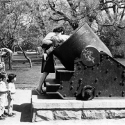 A woman stands near a cannon and boosts a young boy into the muzzle. Another woman and two children stand nearby. The cannon is heavily marked with initials.