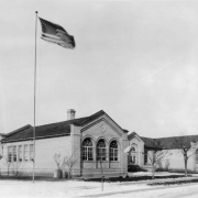 View of Elyria School at 4725 High Street in the Elyria-Swansea neighborhood of Denver, Colorado; the brick building has arched windows and a tile roof. A United States flag waves on a pole.