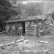 Men pose by a miner's cabin identified as Hepburn's cabin in Miners Basin in the La Sal Mountains (Grand County), Utah. Shows mining tools, ropes and a rifle. The log cabin has a stone chimney and a sod roof.
