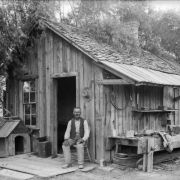 Hughy, a man in cowboy boots and a vest, holds a pipe and sits in front of a wood cabin with a wooden shingle roof in Colorado or Utah. A wooden doghouse is by the porch. Metal buckets, a branding iron, a drill bit, bundles of wire, hats, horseshoes, and a workbench are near the cabin.