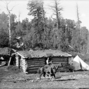 Two men on horseback and a third man stand in front of Geyser cabin in the La Sal Mountains, probably San Juan County, Utah. Signs read, "Geyser cabin," "La Sal and Geyser Express Office," and "Stage Office." Shows a tent beside the cabin and a carcass hanging from a corner of the cabin.