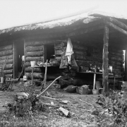 A boy sits in front of Buckeye Cabin in the La Sal Mountains, Utah. A sign reads, "Assay Office Buckeye Cabin." On a table are reagent bottles, a box of assay weights, a beaker, test tubes, glass-stoppered acid bottles, evaporating dishes, and a hot air sterilizer. Also shows a rifle, a shotgun, a gold washing pan, a pickaxe, a hand pick, and buckets.