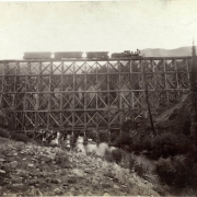 A Denver and Rio Grande narrow gauge locomotive stops on a high wooden trestle over the Lake Fork of the Gunnison River near Lake Fork Junction, Gunnison County, Colorado. Workmen look out from the engine cabin. Shows boulders and brush.