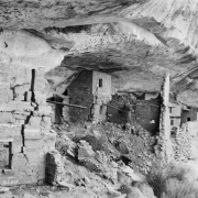View of Native American (Anasazi) cliff dwelling ruins at Balcony House in Mesa Verde National Park, Colorado. Shows multi-story buildings with windows and stone sills beneath a rock wall. A man, probably a guide, stands near a viga.