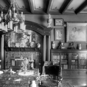 Interior view of the Alonzo Thompson residence (Baerresen Brothers Architects) in Denver, Colorado; decor includes a fireplace, carved wood columns, a chandelier, urns, bookshelves, china, framed paintings, a coffered ceiling, and stencils. A table and chair have acanthus motifs; a mosaic depicts a woman at a spinning wheel.