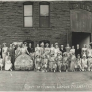 Panoramic view of men and women actors in the Junior League Follies presented by the Junior League of Denver; they pose outdoors in Denver, Colorado. The women wear costumes that include dresses, hats, feather headdresses, and cowgirl outfits. The men wear ties and sweaters, tuxedoes, and two men are dressed as women. A rusticated stone building with arched window lights is nearby.