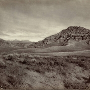 Panoramic view of sedimentary rock formations identified as "Badlands", probably in Fremont County, Wyoming. Shows layered strata on steep rock hillsides. Sagebrush and grass are on sandy, barren ground.