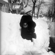 A woman stands in a snow drift from the great snowstorm of 1913 on Clarkson Street in Denver, Colorado. Shows a fur muffler, a possibly Persian lamb's wool coat with fur trim, leather pocketbook and wool hat.