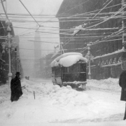 Pedestrians trudge through snow on Welton and Sixteenth (16th) streets in the snowstorm of 1913 in Denver, Colorado. Shows a Denver City Tramway trolley car and snow-laden electrical wires, lamp posts and buildings. The Daniels and Fisher building is in the distance.