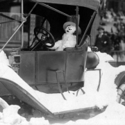 A snowman wears a hat as it sits in the driver's seat of probably a Model T Ford after the snowstorm of 1913 in Denver, Colorado. Pedestrians walk down sidewalk near piles of snow.