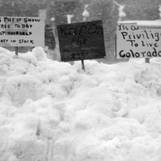 View of signs stuck in a snow pile from the 1913 blizzard in Denver, Colorado. Signs read, "This pile of snow free to day help yourself plenty in stock," "Keep Off the Grass," "It's a Privilige [sic] To Live in Colorado."