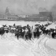 Men in Civic Center unload snow laden horse-drawn wagons after the snowstorm of 1913 in Denver, Colorado. Shows pedestrians on the sidewalk, the Arapahoe County Courthouse building and the Majestic Hotel are in the distance. A sign reads, "Oil and Gas (?) 500,000 Population for Denver, the Colorado Producers Oil Company."