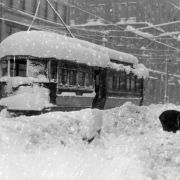 A snow covered Denver City Tramway trolley car is stuck in the snowstorm of 1913 in central Denver, Colorado. Signs on a building read, ""Oriental Rugs," and "Store Room For Rent, 20 x 40 ft." A man makes a large snowball.