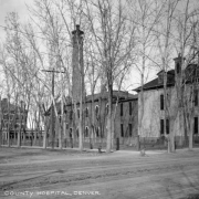View of several brick structures associated with Arapahoe County Hospital (Denver General Hospital) at West 6th (Sixth) Avenue and Cherokee Street in Denver, Colorado. Shows the Training School building with two-tier porch in the distance. The chimney from the plant building extends above the trees.