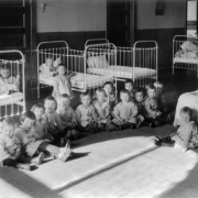 Orphans at the Denver Orphans' Home (later the Denver Children's Home) at 1501 Albion Street in the South Park Hill neighborhood of Denver, Colorado sit on the floor and in cribs in their dormitory and pose for the camera. A woman caregiver looks at the children.
