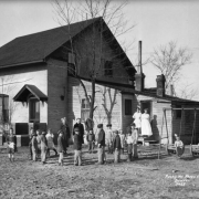 Boys and girls stand in a circle and pose in the backyard of a small house used as an orphanage while the Denver Orphans' Home is constructed. Shows children in swings on a swingset. Three women caregivers stand with the children.