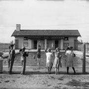 Children stand behind a wire fence and watch a boy in a bathing suit prepare to dive into the Children's Pool at Elyria Playgrounds (Elyria Park) at 48th and High Streets in the Elyria Swansea neighborhood of Denver, Colorado. Shows a stucco building with tile roof and brick quoins. An adult in a swimsuit watches the diver; other swimmers stand in the water and sit at the pool's edge.