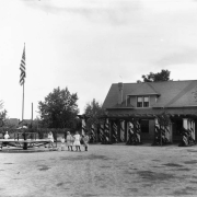 Children pose next to the merry-go-round and flagpole at the Elyria Community Center in Elyria Park, 4809 High Street in the Elyria Swansea neighborhood of Denver, Colorado. Shows a two story brick building with a dormer and a stone column pergola.
