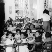 Two African American (Black) women serve food to African American and white boys and girls at the George Washington Carver Day Nursery at 2260 Humboldt Street in the City Park West neighborhood of Denver, Colorado. Shows a room with a high ceiling and tall windows framed by curtains. The children sit at low, long tables; some drink milk and look back toward the camera.
