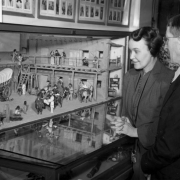 A man and woman examine an exhibit at the Colorado Museum of Natural History in Denver, Colorado. A diorama features a covered wagon, people, horses, and an adobe building with vigas and a balcony.