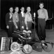 Group portrait of musicians in Denver, Colorado who pose indoors by an accordion, violin, bass viola, steel and acoustic guitars, a banjo, and a microphone with letters: "NBC." Men's outfits include western shirts, bandanas, and cowboy boots.