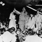 Jeannette Lynn is crowned the queen of the fiftieth anniversary celebration of the town of Edgewater, Jefferson County, Colorado. She and Mayor Lloyd Doherty stand in the center of a stage; on the left is Ruth Dillner and on the right is Betty Gottschalk, who were attendants; the three women wear strapless evening gowns. Several men in suits and two women, one eldery, are also on the stage. A crowd looks up at the ceremony.