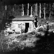 Two men stand on a grassy slope outside a log cabin with a tar paper roof near the mining communty of Hahn's Peak (Routt County; also known as Poverty Bar), Colorado.  Piles of boards litter the ground behind the cabin.