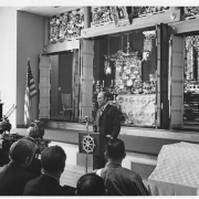 A black and white photograph of Sterling Kahn speaking to a group at the Buddhist temple.  He is standing at a podium in front of the altar.
