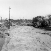 View of a Cherry Creek flood in Denver, Colorado after the Castlewood Canyon Dam break; shows torrents of muddy water. West Denver High School is to the left.