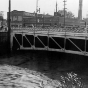 View of a Cherry Creek flood in Denver, Colorado after the Castlewood Canyon Dam break; shows the Broadway Street bridge, people, and a car dealership with a smokestack and signs: "Continental, Buick."