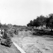 View of a Cherry Creek flood in Denver, Colorado after the Castlewood Canyon Dam break; shows torrents of muddy water in standing waves. People look on from the side.
