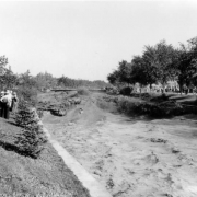View of a Cherry Creek flood in Denver, Colorado after the Castlewood Canyon Dam break; shows torrents of muddy water in standing waves. People look on from the side; boys wear plastic visors.