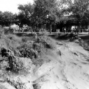 View of a Cherry Creek flood in Denver, Colorado after the Castlewood Canyon Dam break; shows torrents of muddy water in standing waves. People watch from under trees.