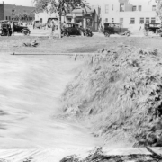 View of a Cherry Creek flood at 12th (Twelfth) Avenue in Denver, Colorado after the Castlewood Canyon Dam break; shows torrents of muddy water, parked cars, and people.