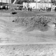 View of a Cherry Creek flood between 12th (Twelfth) and 13th (Thirteenth) Avenues in Denver, Colorado after the Castlewood Canyon Dam break; shows torrents of muddy water, standing waves, parked cars, and people.
