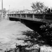 View of a Cherry Creek flood in Denver, Colorado after the Castlewood Canyon Dam break. People stand on the 13th (Thirteenth) Avenue bridge and watch torrents of water.