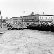 View of a Cherry Creek flood in Denver, Colorado after the Castlewood Canyon Dam break; shows muddy water, the Blake Street bridge, people, debris, and warehouses with signs: "Mile High Poultry Farm."